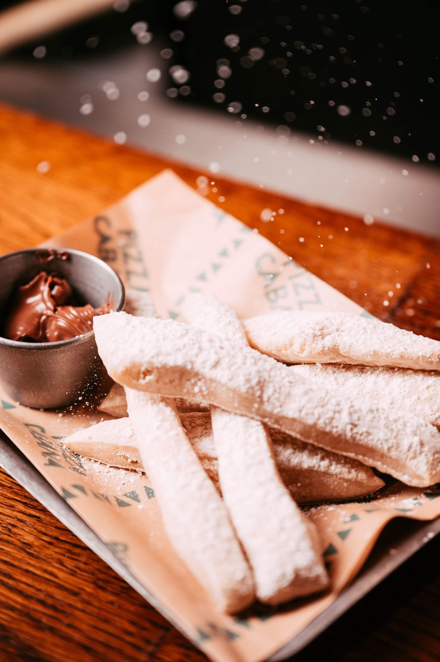 Image of sliced up dough sticks with a pot of nutella next to them, being sprinkled in icing sugar on a wooden board. 