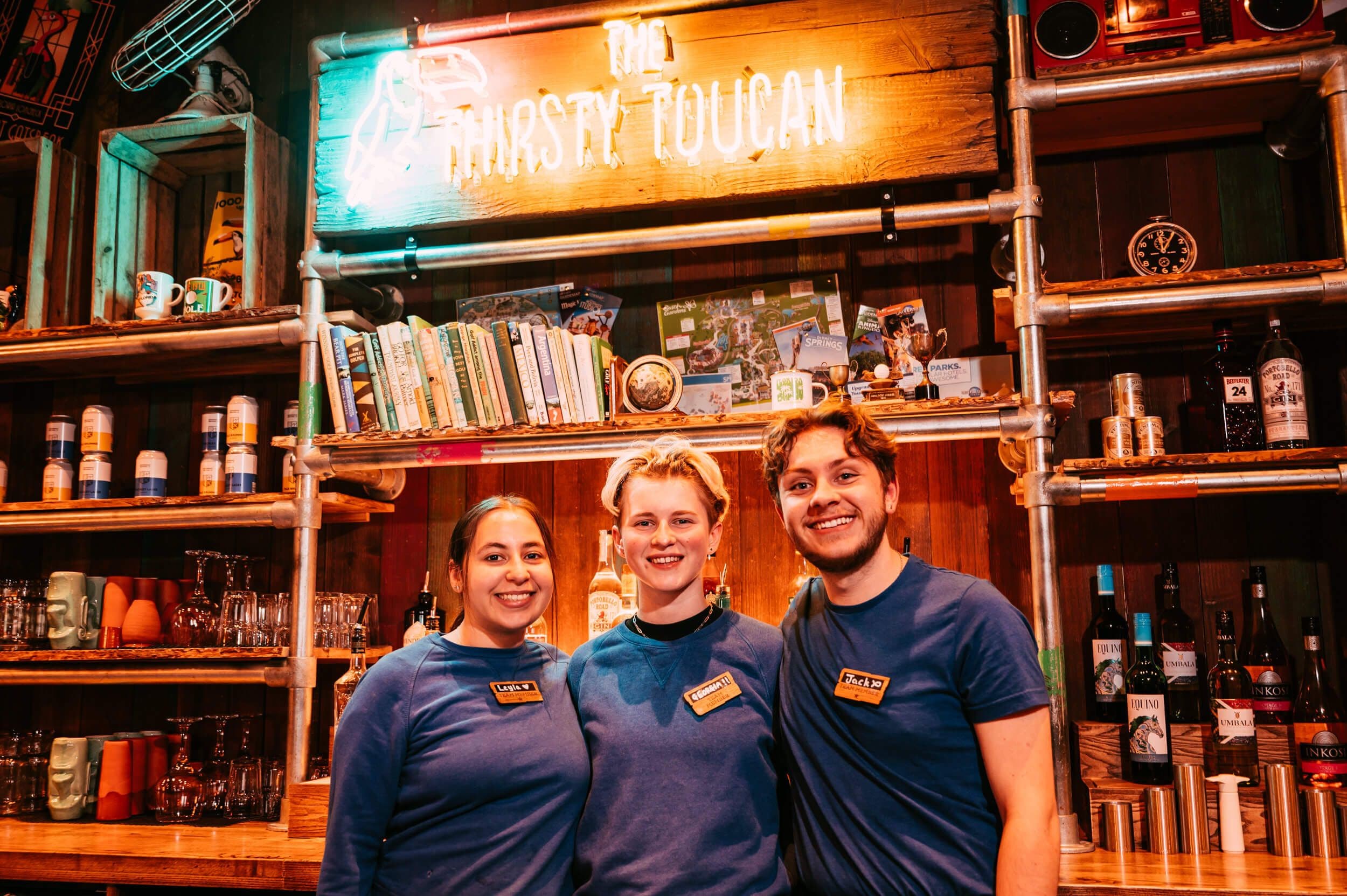 Tree members of the team of the Thirsty Toucan, standing behind the bar wearing blue shirts and smiling. 