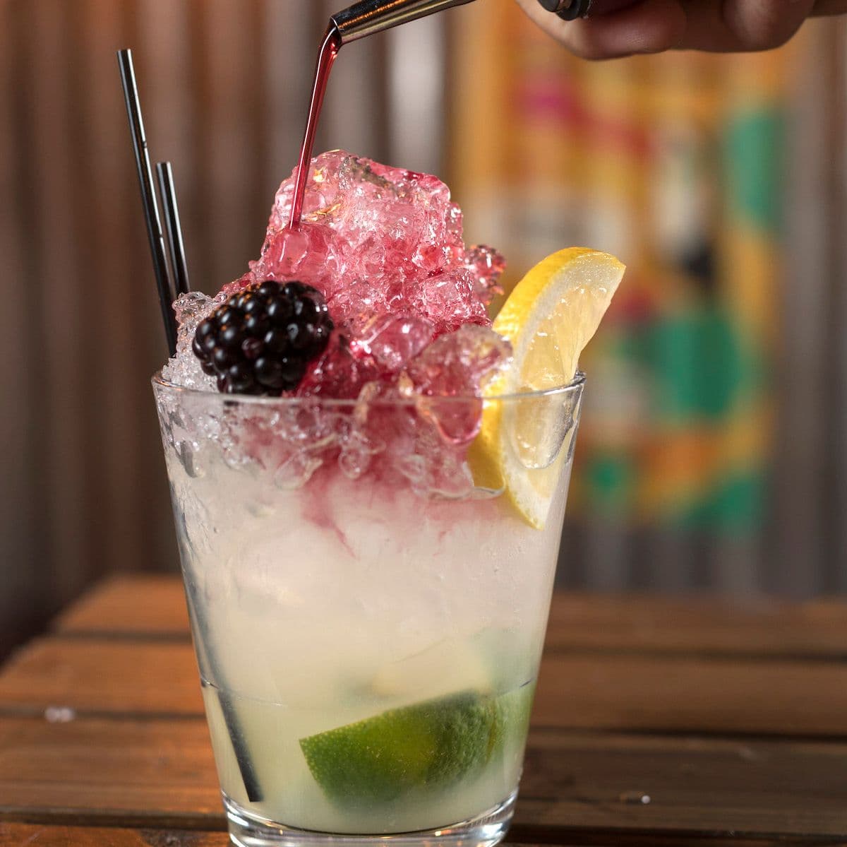 Image of delicious blackberry liquor being poured over a Samba Bramble cocktail, with fresh lemon, lime and blackberry garnish.