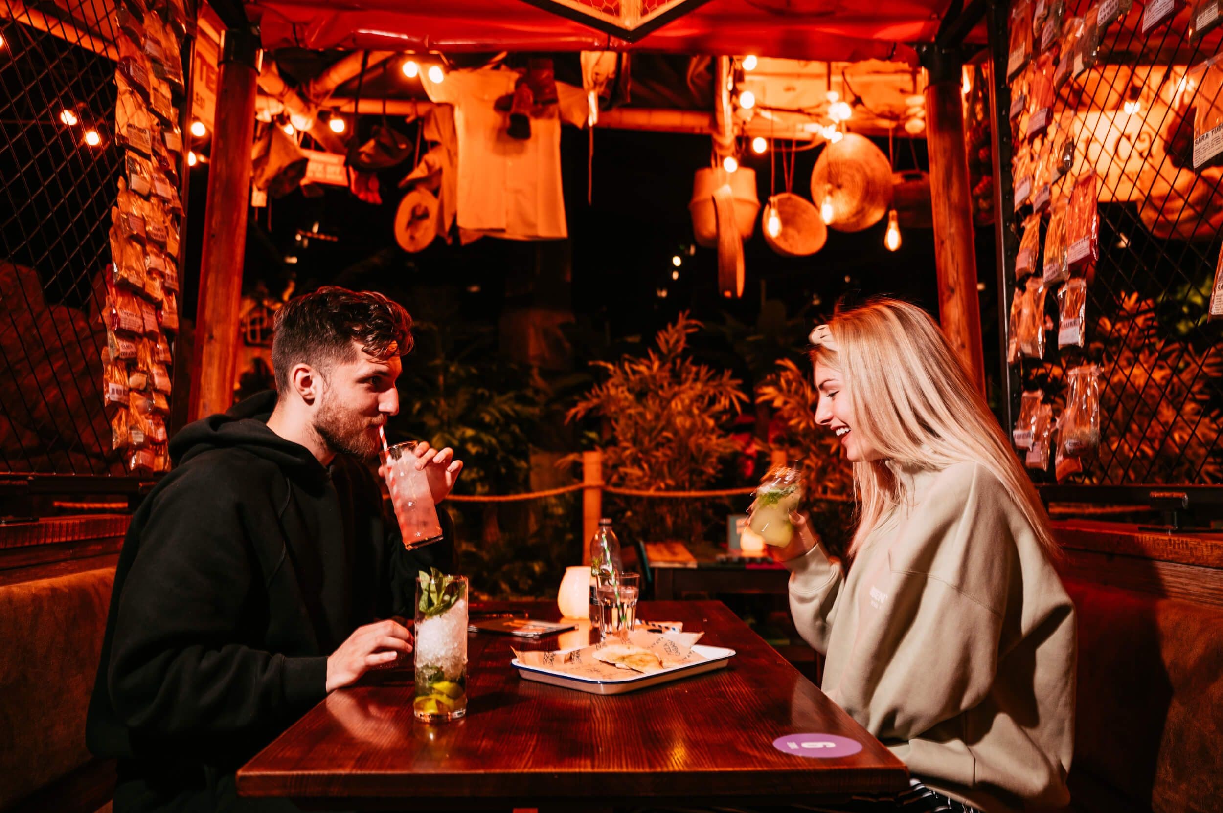 A couple sit enjoying some Thirsty Toucan Bar cocktails in the Treetop market, with glowing lanterns around them.