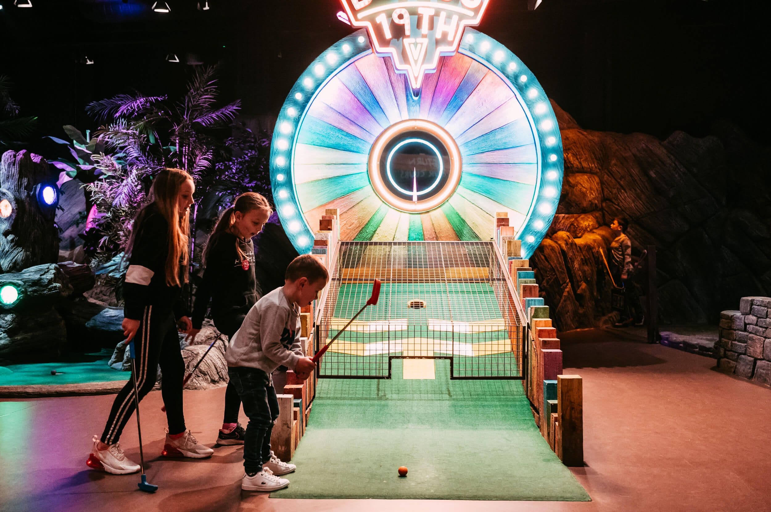 Three young children shoot their shot at the Bonus 19th Hole spin wheel, with bright lights shrouding them from every angle.